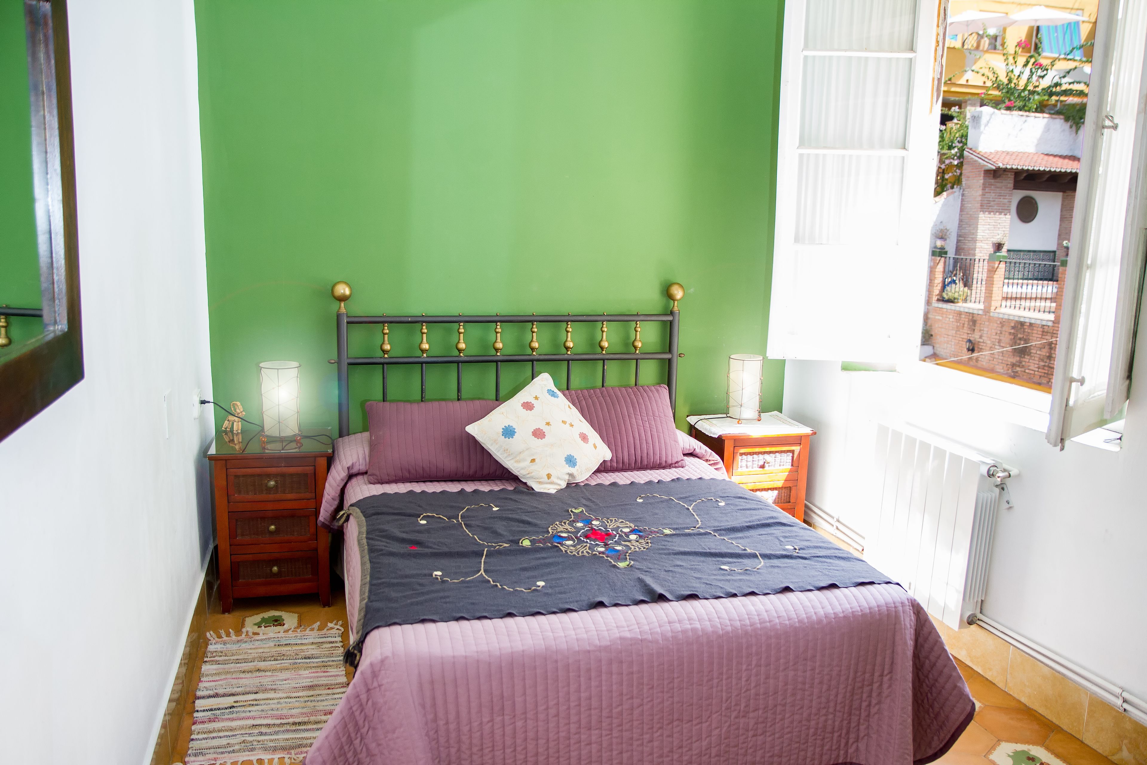 Une chambre aux murs verts, avec un lit recouvert d’une couverture violette, une table de chevet en bois et une fenêtre donnant sur un balcon.