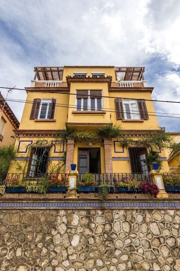 A two-story building with a yellow facade, a stone base, and a balcony with a wooden railing.
