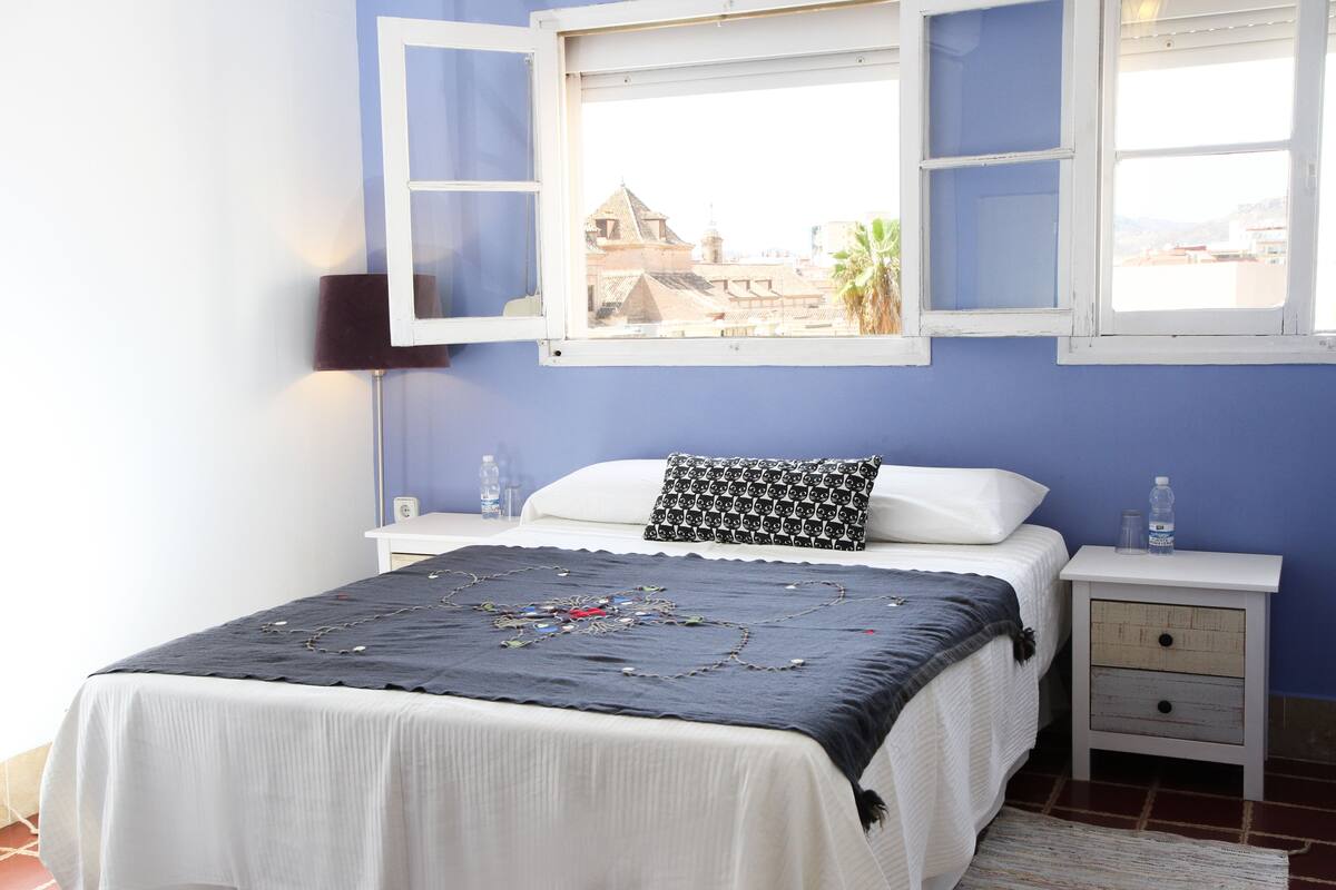 Une chambre à coucher avec un lit, une table de chevet, une fenêtre et une vue sur les bâtiments à l’extérieur.