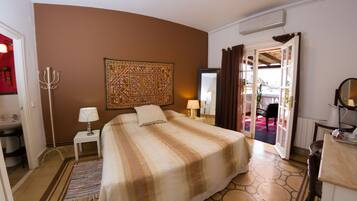 Habitación de hotel con una cama grande, una mesita de noche pequeña con lámpara, un adorno en la pared y vistas al exterior a través de una puerta abierta.