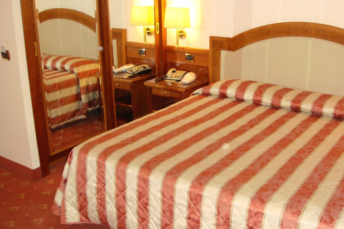 Una habitación de hotel con una cama grande, una mesita de noche con lámpara, un teléfono y un espejo.