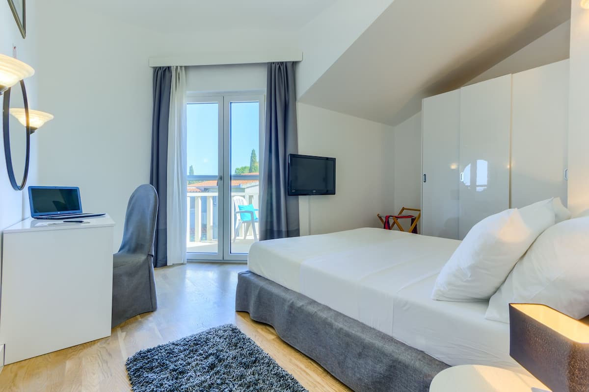 A modern hotel room with a bed, a desk with a laptop, a TV, and a balcony with a view.