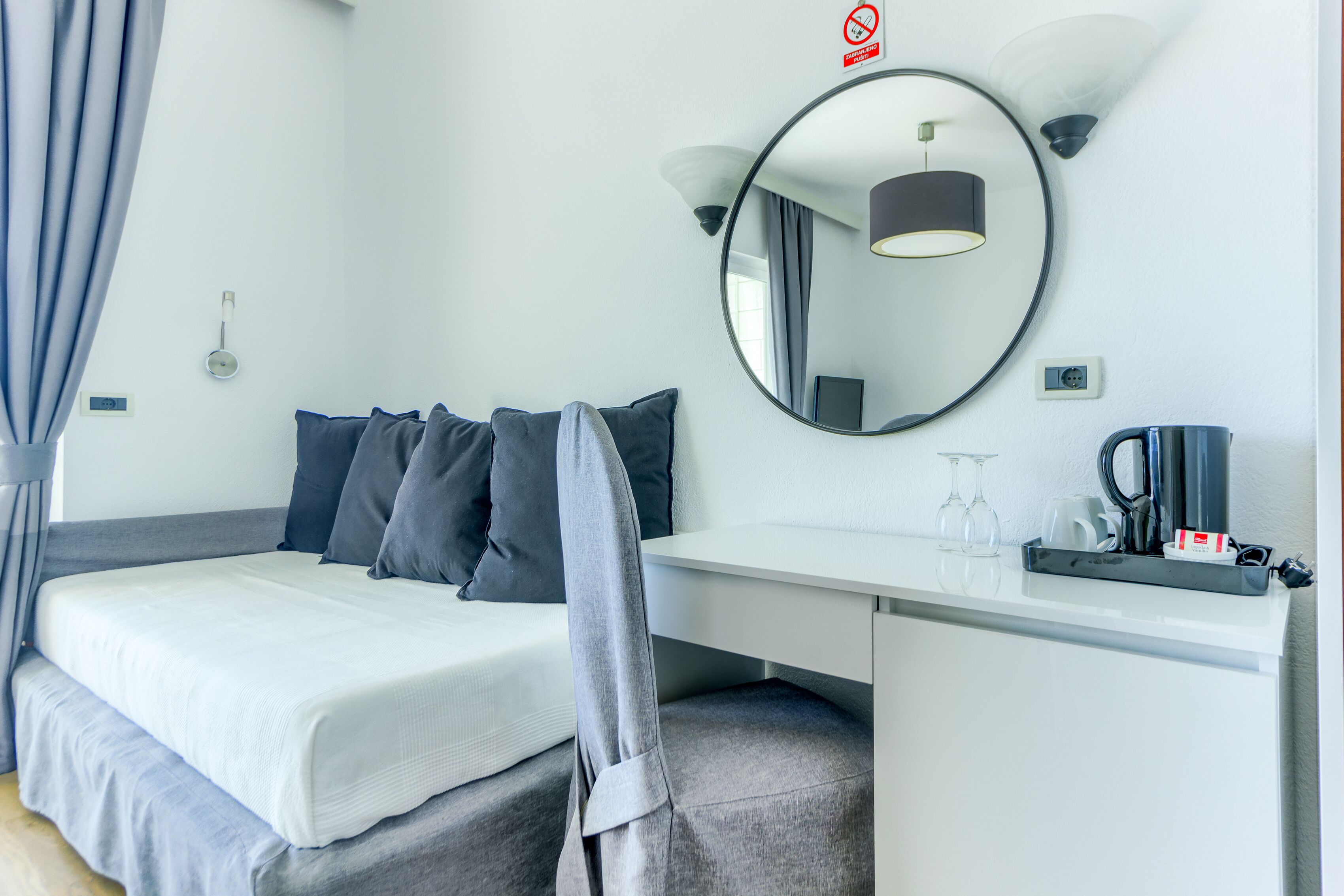 A hotel room with a bed, a desk, a mirror, a coffee maker, and a small table with a glass and a cup.