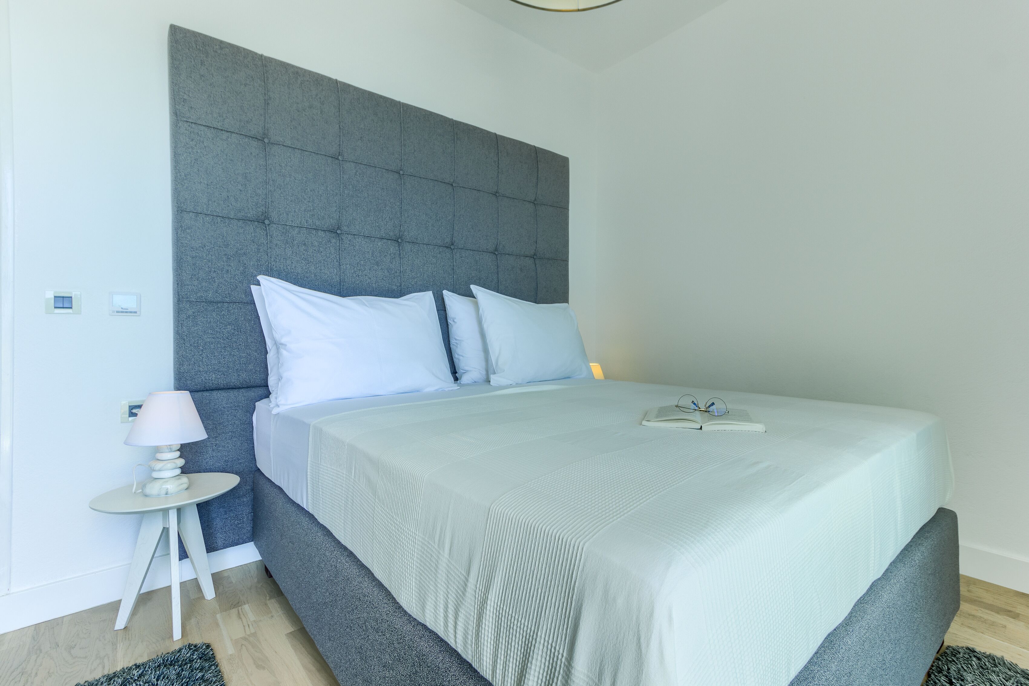 A modern bedroom with a grey upholstered headboard, a neatly made bed with white linens, and a small bedside table with a lamp.