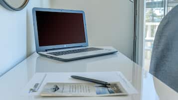 A laptop on a white table with a pen and papers, near a window with a view of trees.