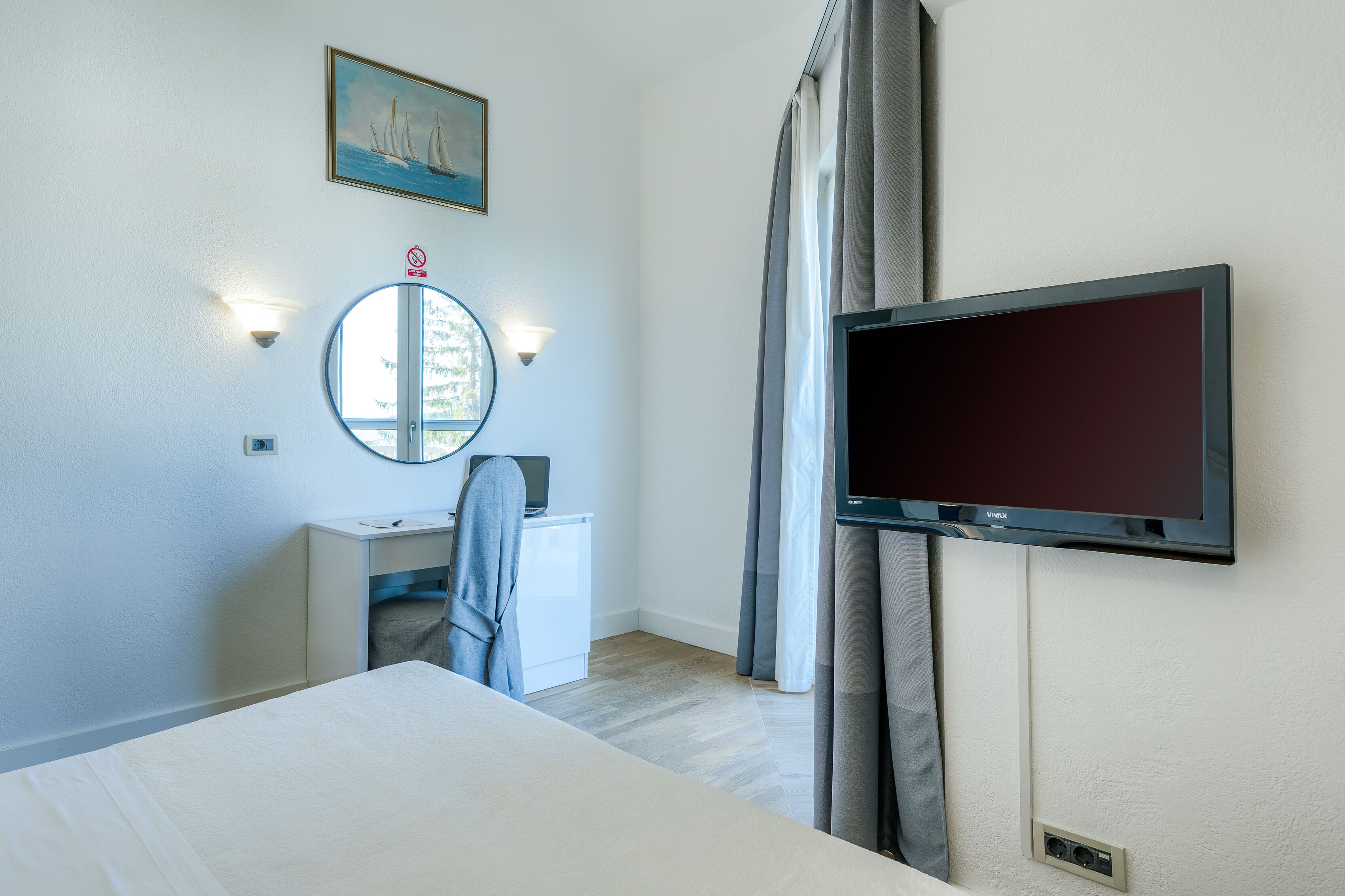 A hotel room with a bed, a desk with a chair, a TV mounted on the wall, a round mirror, and a painting above the mirror.