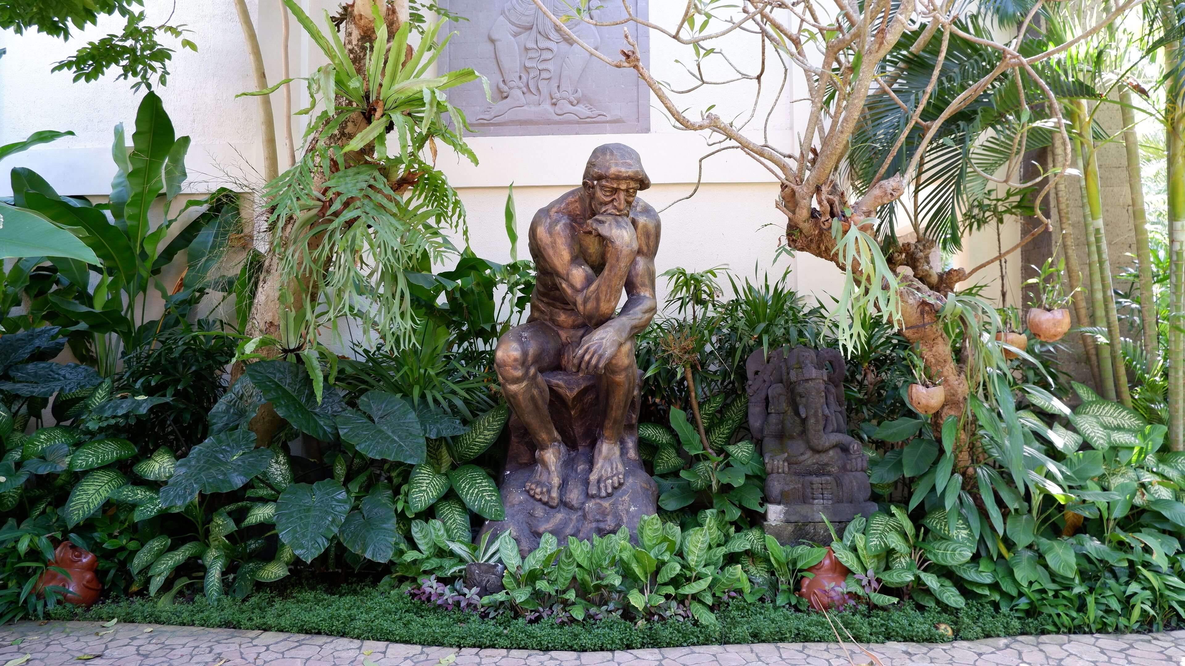 Una statua di un uomo seduto, circondata da rigogliosa vegetazione tropicale e un rilievo in pietra sulla parete.