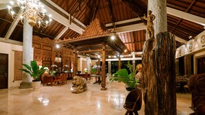 A spacious lobby with a high ceiling, ornate wooden ceiling, a large central column, and a seating area with chairs and a table. - Hotel Kumala Pantai (Legian)