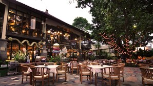An outdoor dining area with wooden tables and chairs, surrounded by a building with large windows and decorative lighting. - Hotel Kumala Pantai (Legian)
