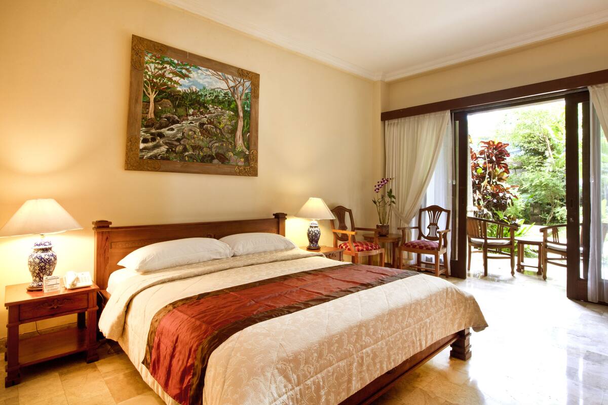 A hotel room with a large bed, two bedside tables with lamps, a painting on the wall, and a view of the garden through the door.