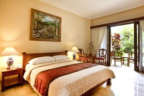 A hotel room with a large bed, two bedside tables with lamps, a painting on the wall, and a view of the garden through the door. - Hotel Kumala Pantai (Legian)