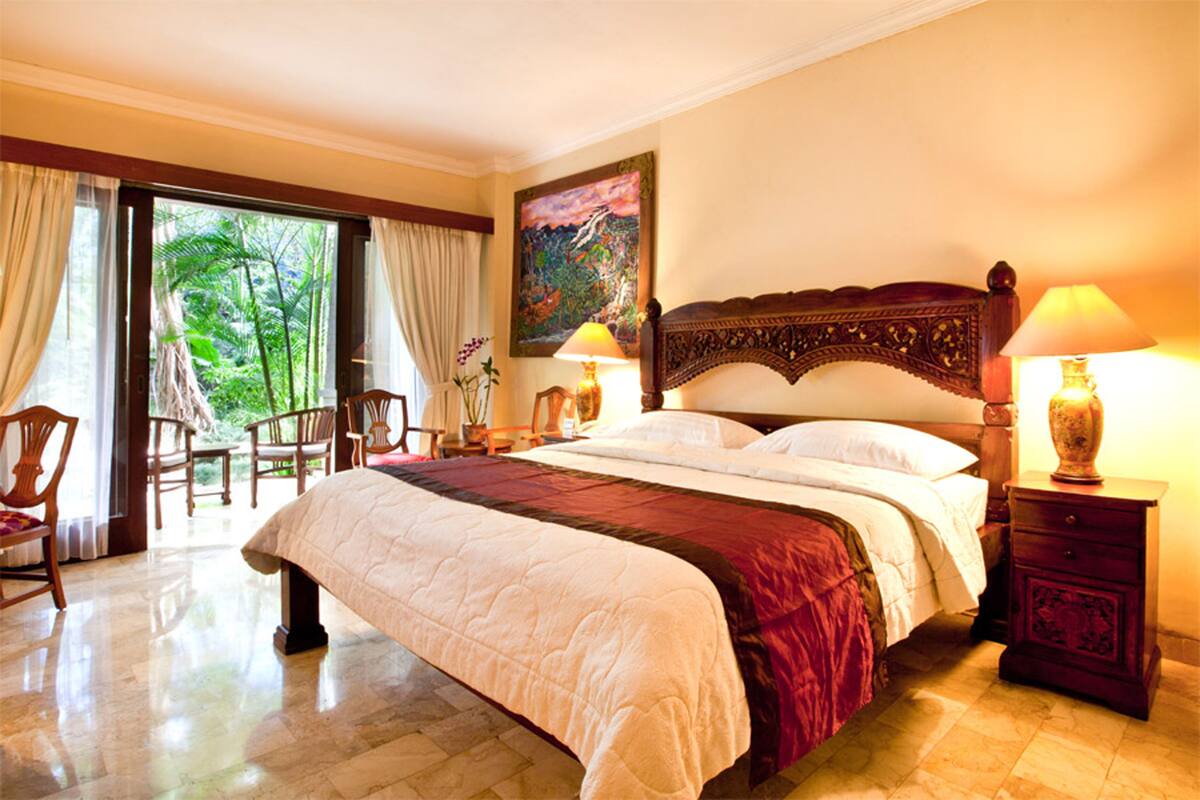 A spacious bedroom with a large bed, a wooden headboard, and a view of lush greenery through a sliding door.