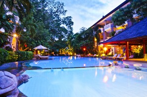 A swimming pool with lounge chairs and umbrellas, surrounded by lush greenery and a building with a thatched roof. - Hotel Kumala Pantai (Legian)