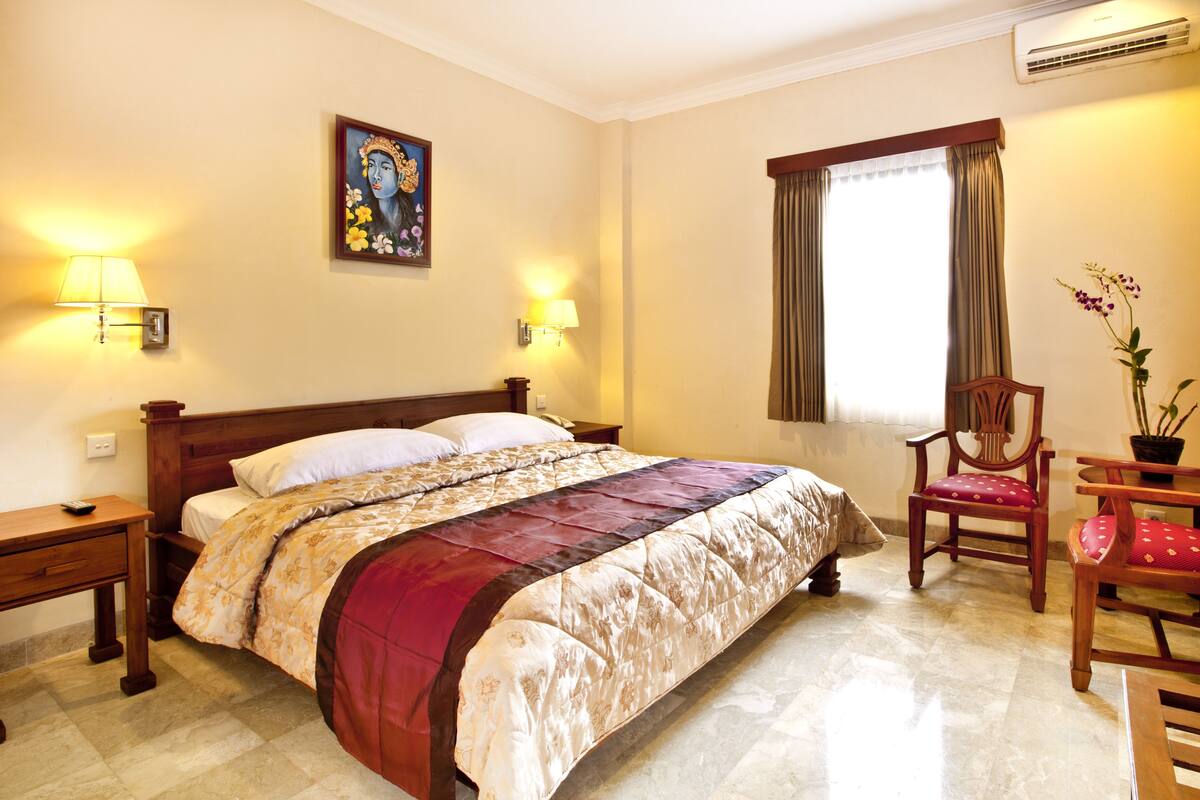 A hotel room with a large bed, two bedside lamps, a wooden chair, a small table, and a window with curtains.