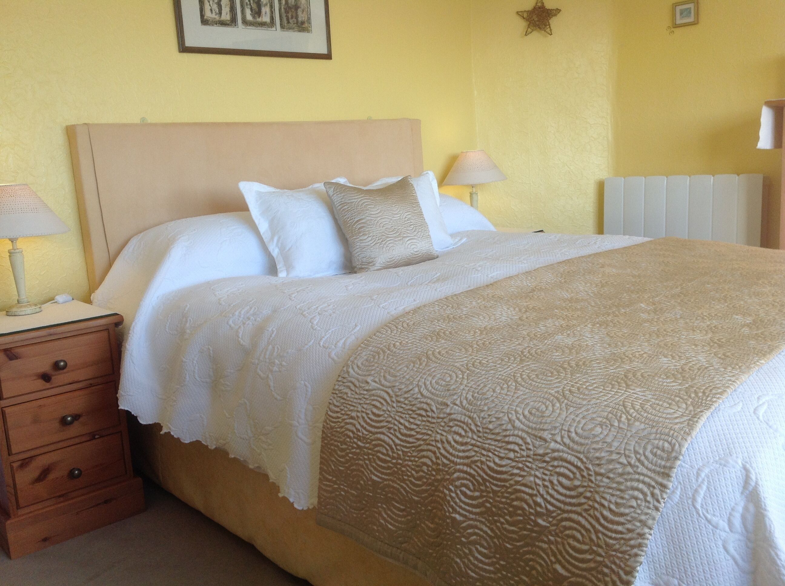 A neatly made bed with a beige headboard, white bedding, and a patterned throw blanket. There is a wooden nightstand with a lamp and a drawer.
