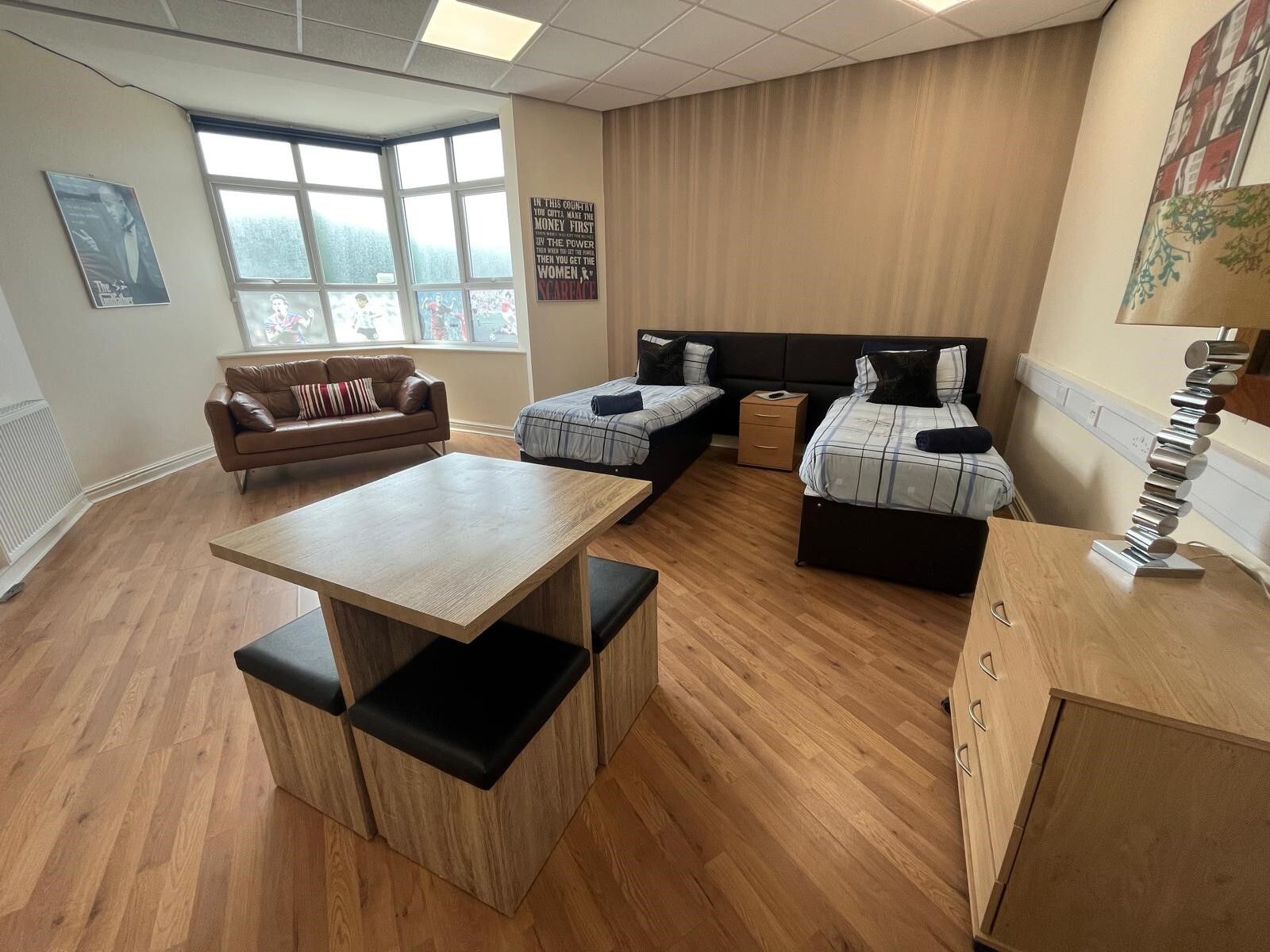 A modern room with a wooden floor, a sofa, two beds with pillows, a small table, and a lamp.