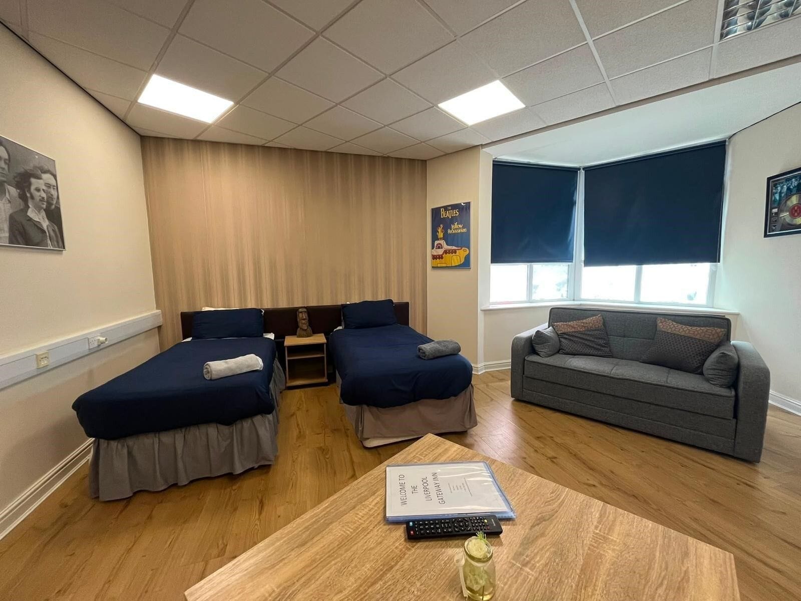 A hotel room with two single beds, a sofa, a small table with a book and remote control, and a poster on the wall.