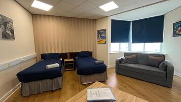 A hotel room with two single beds, a sofa, a small table with a book and remote control, and a poster on the wall.