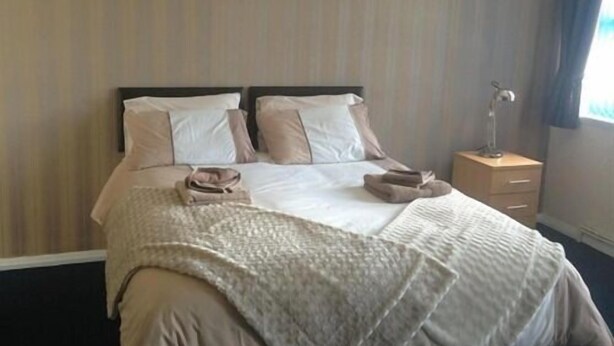 Een netjes opgemaakt bed met een witte deken en twee kussens, een nachtkastje met een lamp, en een raam met gordijnen.