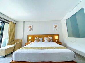 A hotel room with a large bed, a wooden headboard, two bedside lamps, a chair, and artwork on the walls. - Dekuta Hotel (Kuta)