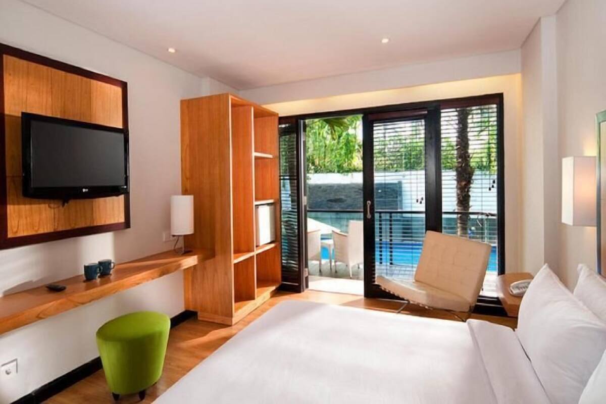 A modern hotel room with a large bed, a TV mounted on the wall, a wooden wardrobe, a green ottoman, and a balcony with a pool view.