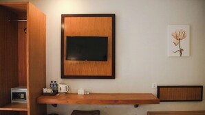A compact room with a wooden desk, a mounted TV, a small stool, and a trash can. - Dekuta Hotel (Kuta)