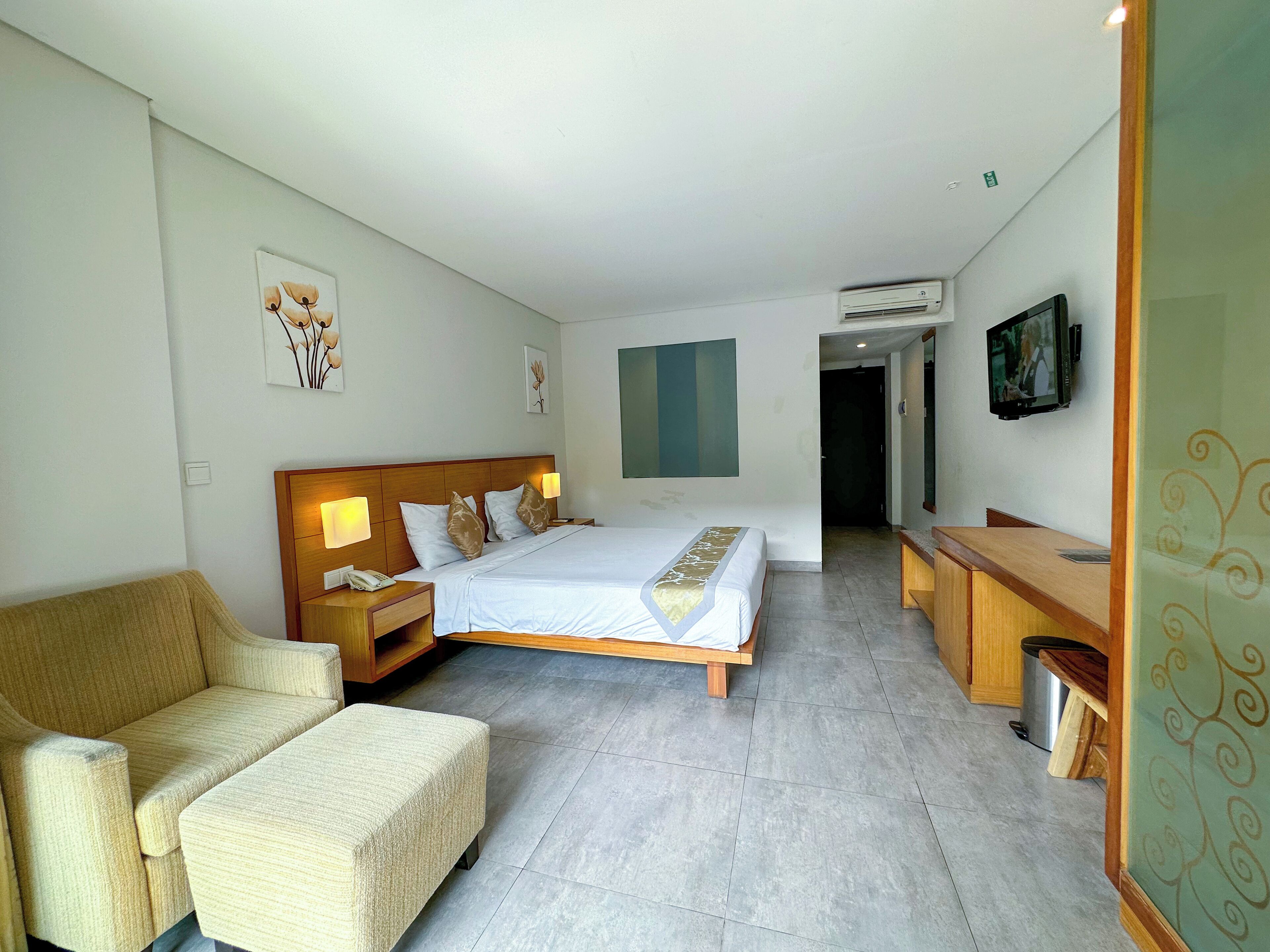 A hotel room with a bed, bedside tables, a chair, a desk, an air conditioning unit, and a television.