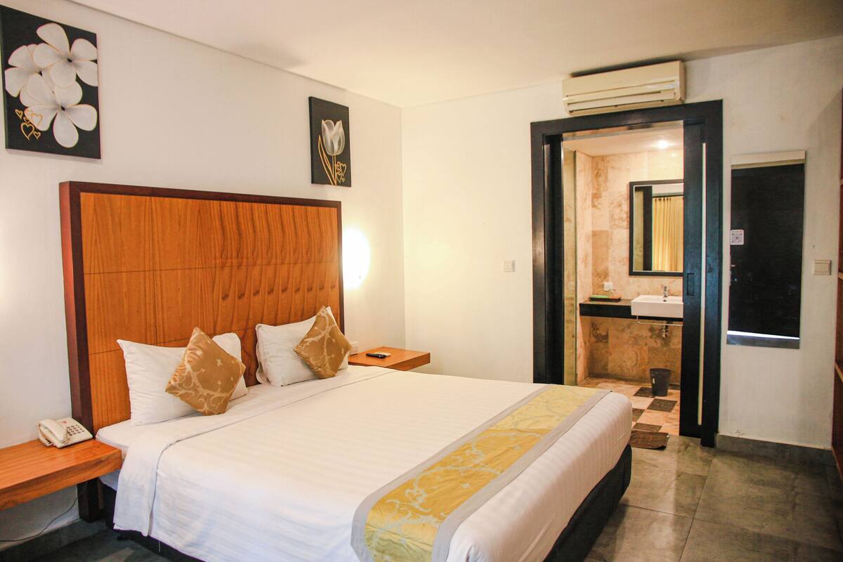 A hotel room with a large bed, wooden headboard, bedside tables, a nightstand, and a bathroom visible through an open door.