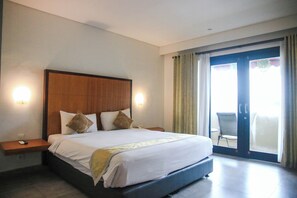 A hotel room with a large bed, two bedside tables, a nightstand, and a view of the outside through a sliding glass door. - Dekuta Hotel (Kuta)