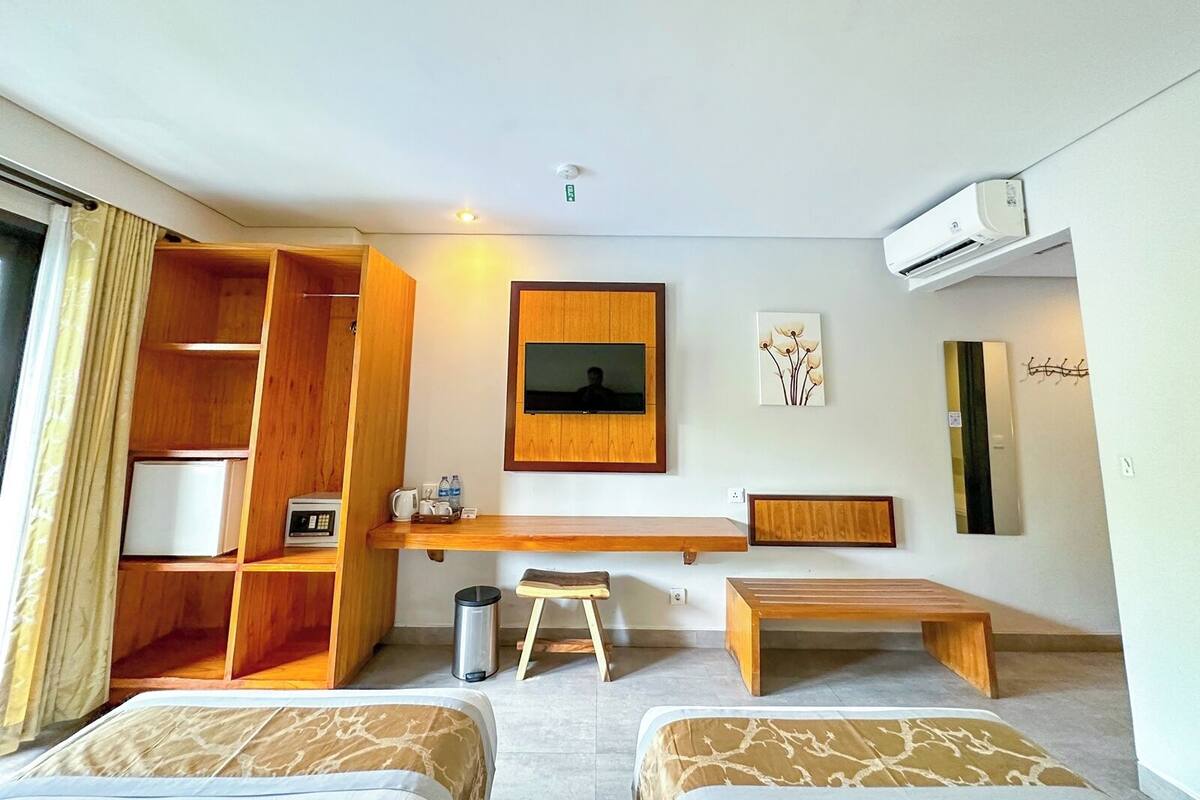 A hotel room with two beds, a wooden desk, a TV, a small stool, and a trash bin.