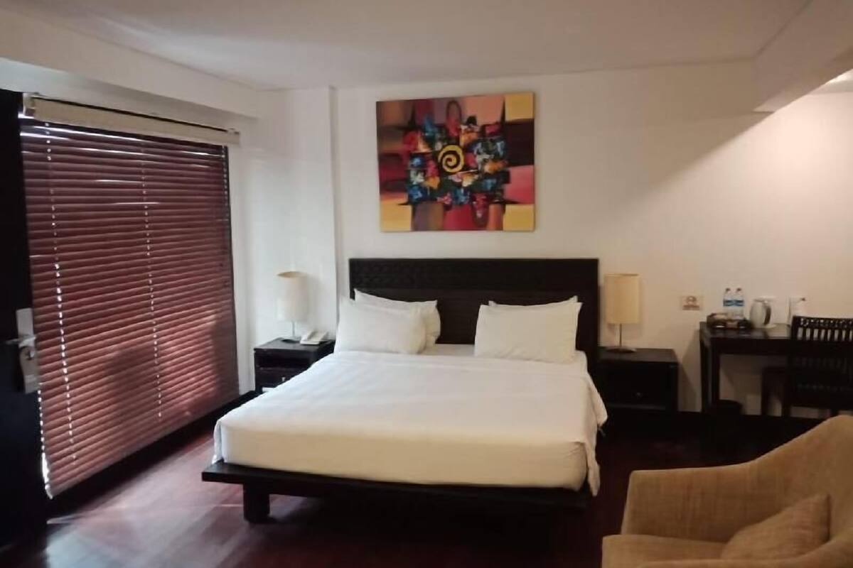 A hotel room with a bed, a desk, a chair, a painting on the wall, and a window with blinds.