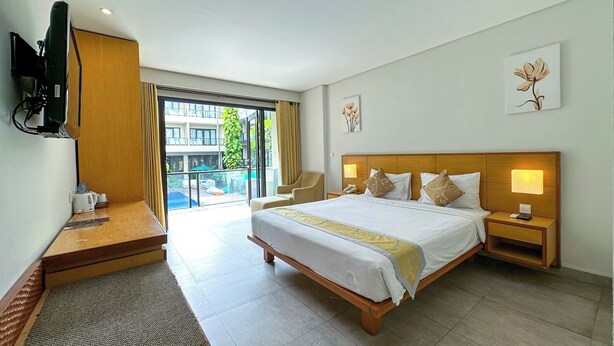 A hotel room with a large bed, bedside tables, a desk, a television, and a balcony with a view of buildings and greenery. - Dekuta Hotel (Kuta)