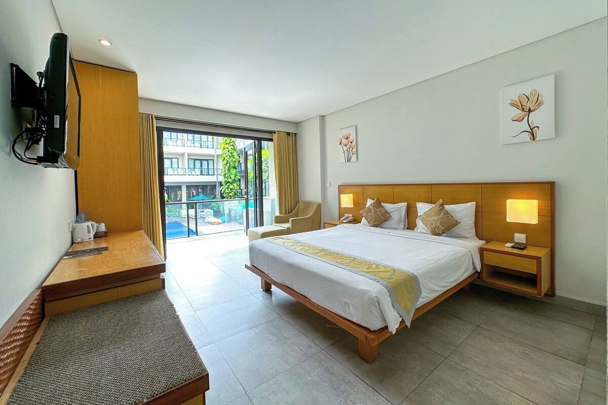 A hotel room with a large bed, bedside tables, a desk, a television, and a balcony with a view of buildings and greenery.