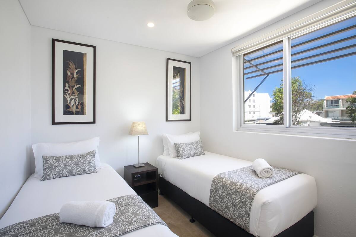 Une chambre d’hôtel avec deux lits, une table de chevet, une lampe et une fenêtre donnant sur des immeubles.