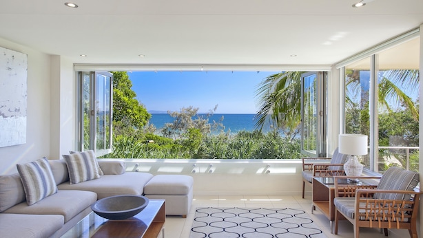 Ein modernes Wohnzimmer mit einem großen Panoramafenster mit Blick auf einen Strand, einer Couch, Sesseln und einem Couchtisch.