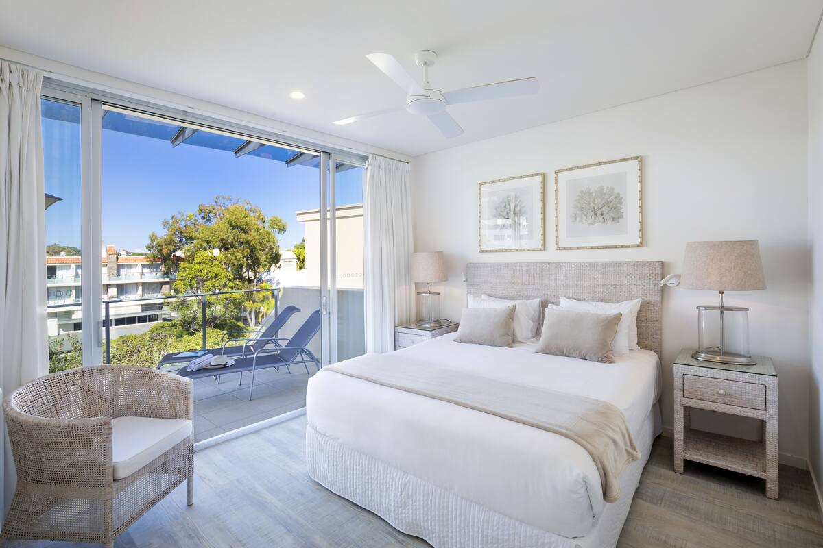 Une chambre spacieuse avec un grand lit, deux tables de chevet, un ventilateur de plafond et un balcon équipé d’une chaise et d’une table.