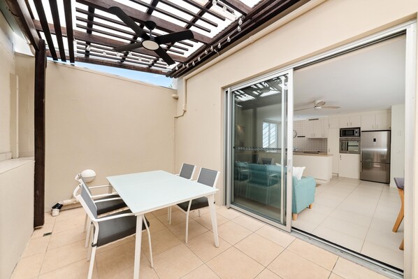 Eine moderne Küche mit Edelstahlgeräten, ein Essbereich mit weißem Tisch und Stühlen sowie eine Glasschiebetür, die auf einen Balkon mit Deckenventilator führt.
