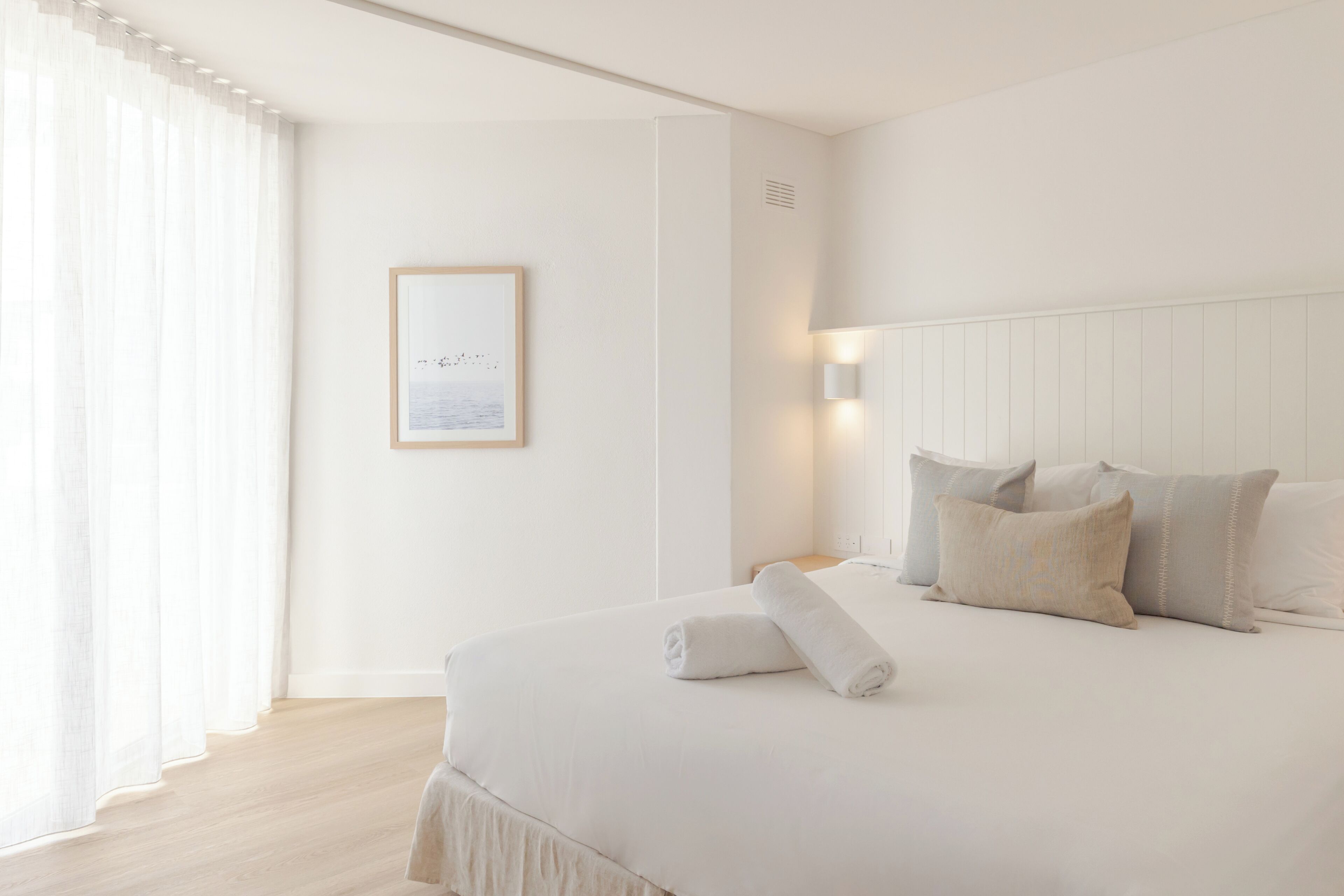 A neatly made bed with a rolled towel, a framed picture on the wall, and a bedside lamp.