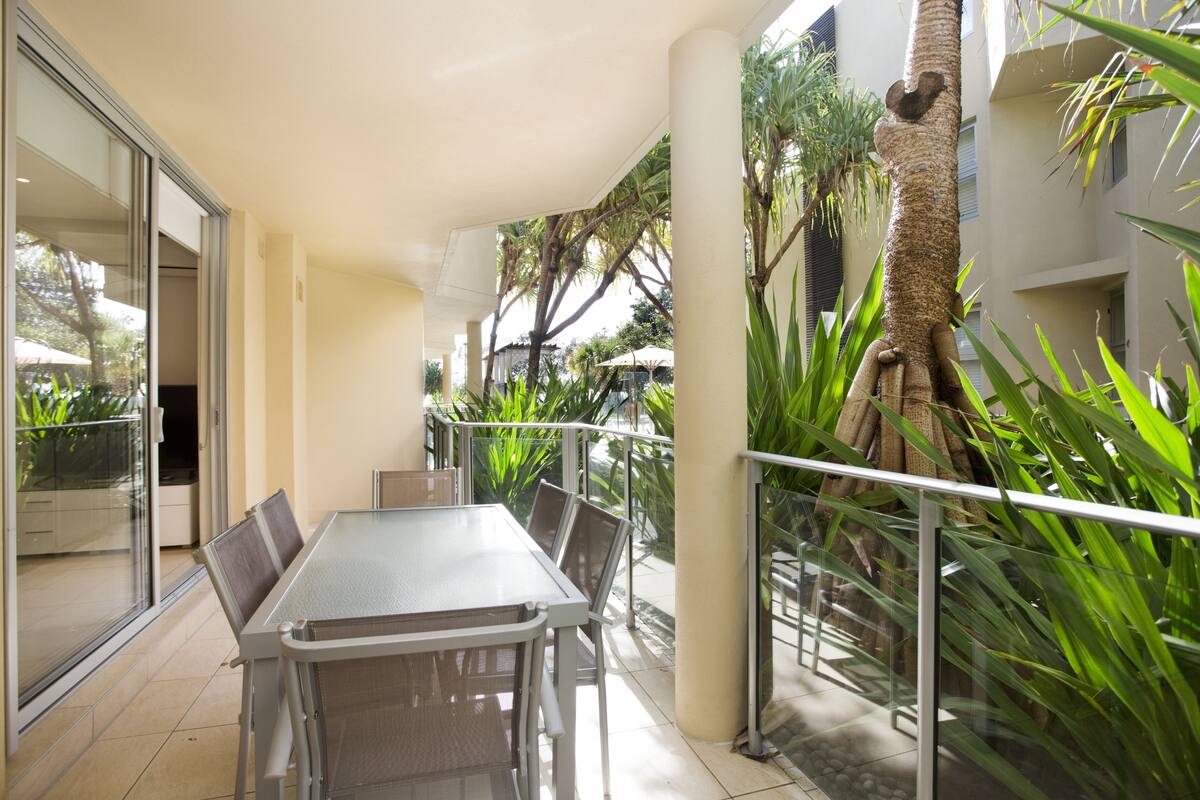 Un balcon avec une porte vitrée donnant sur un patio, une table avec des chaises et un palmier.