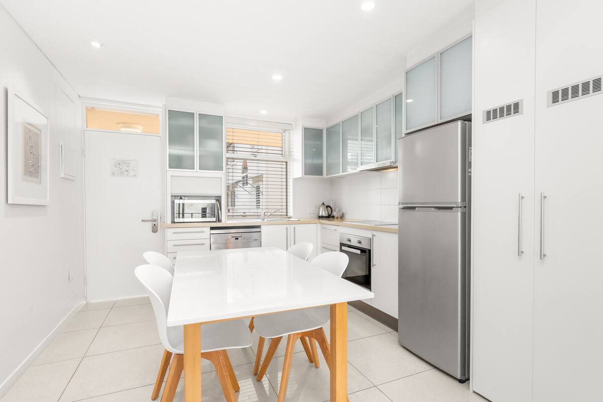 Une cuisine moderne avec des meubles blancs, un réfrigérateur en acier inoxydable et une table à manger blanche avec des chaises.