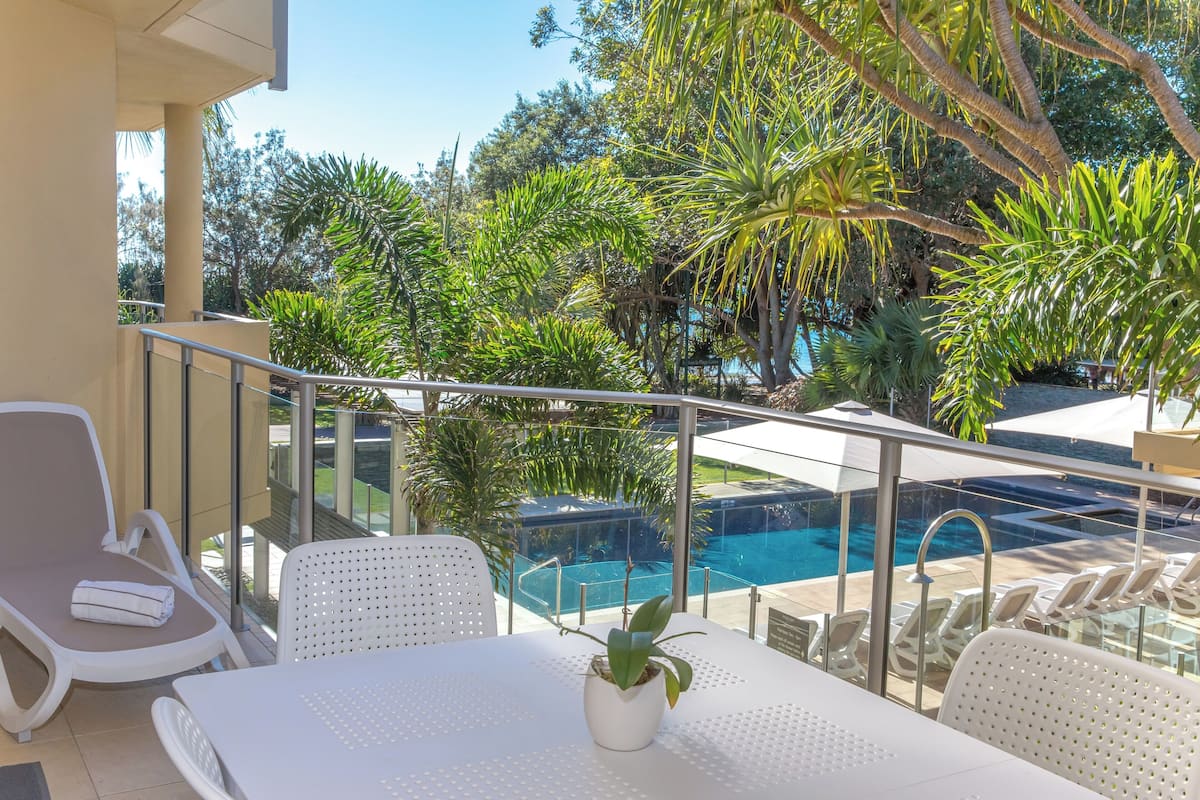 Un balcon avec une table et des chaises, donnant sur une piscine et une végétation luxuriante.