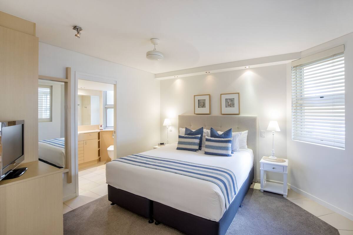 Une chambre moderne avec un grand lit, des tables de chevet, une télévision et une salle de bain visible à travers une porte ouverte.
