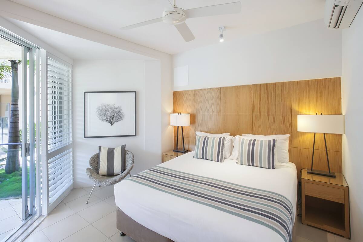 Une chambre d’hôtel moderne avec un grand lit, deux lampes de chevet, un ventilateur de plafond et une vue sur l’extérieur grâce à une porte coulissante.