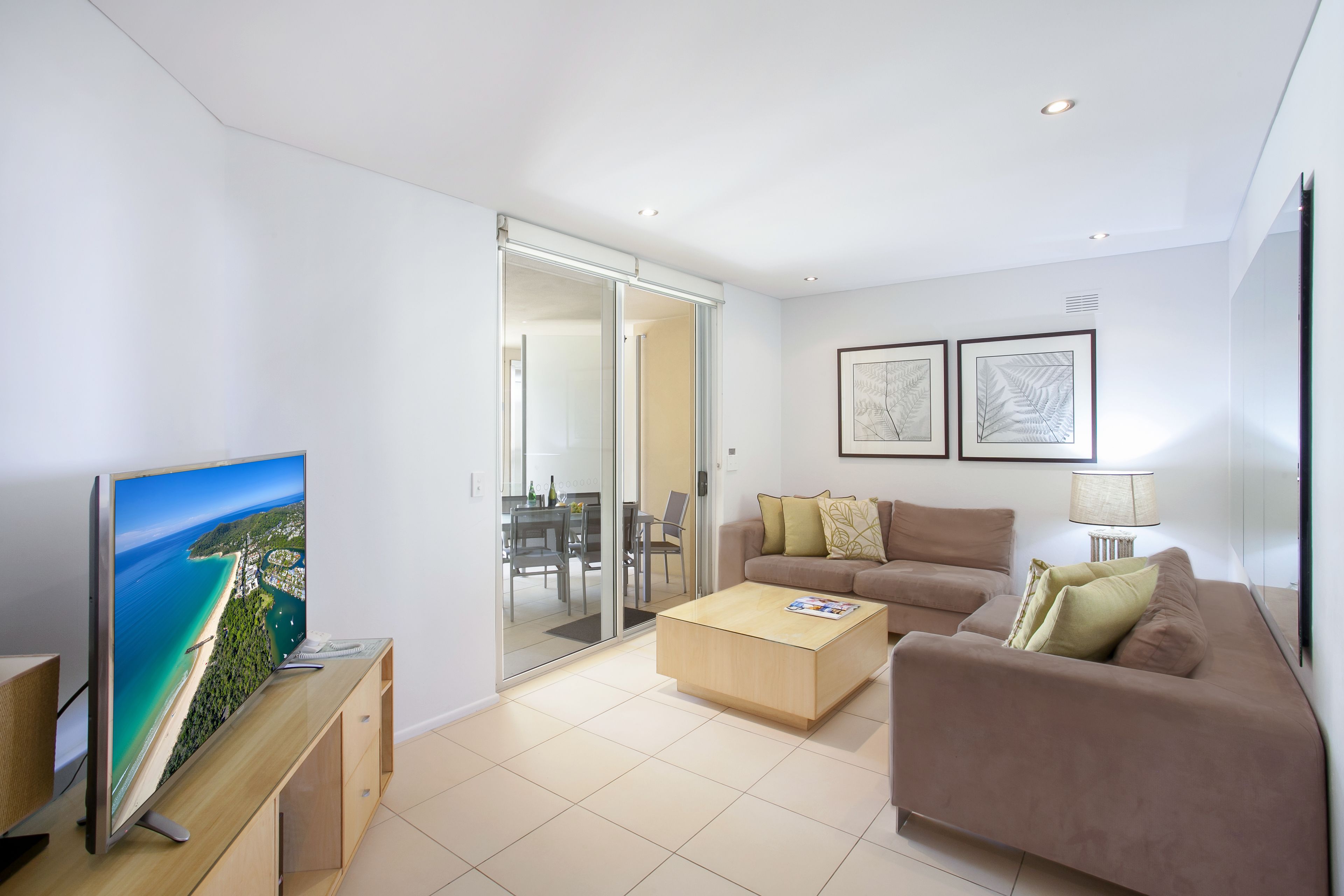 A modern living room with a flat-screen TV, a brown sofa, a wooden coffee table, and a glass door leading to a balcony.