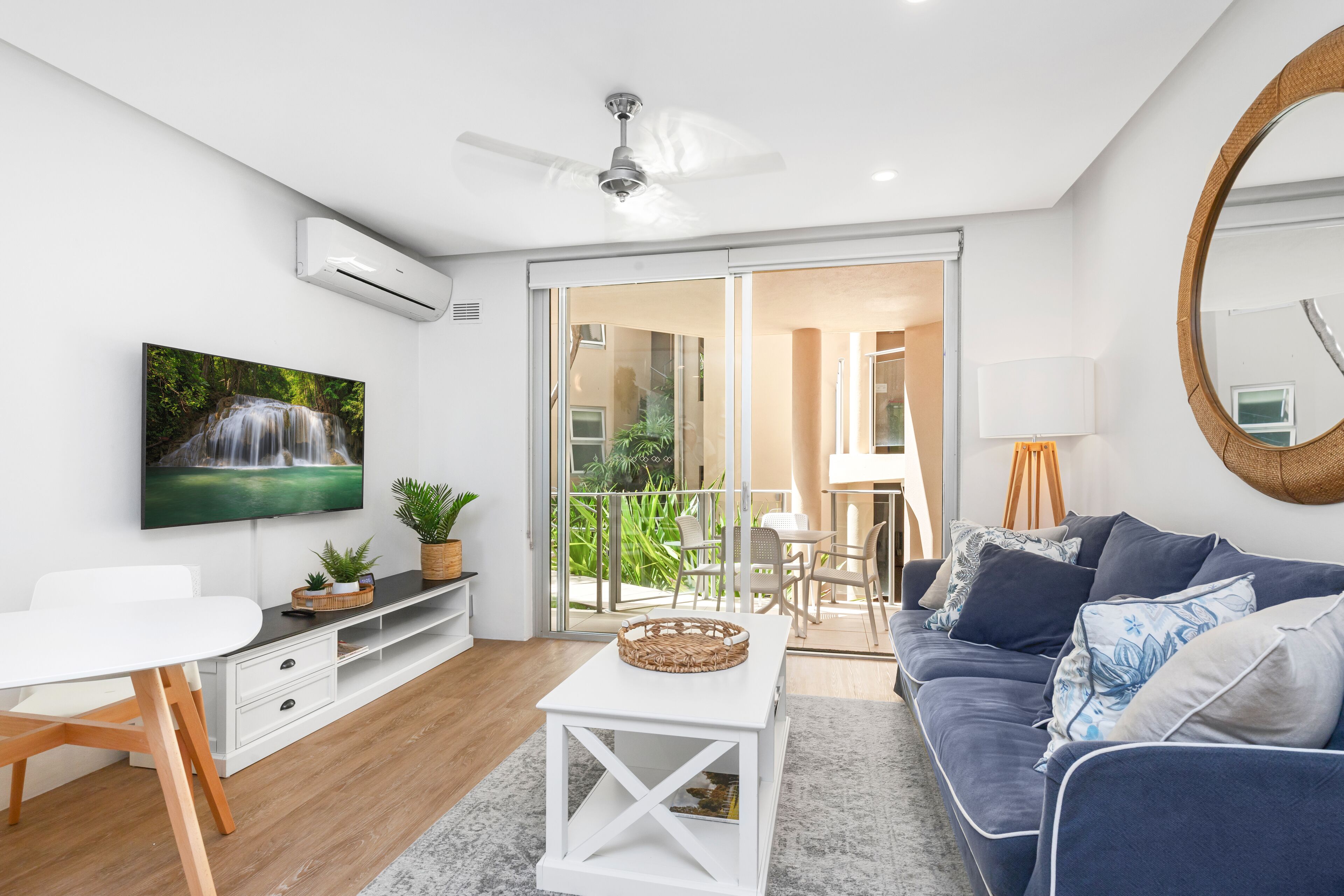 Una sala moderna con televisor de pantalla plana, ventilador de techo, comedor con mesa y sillas, y balcón con vistas a la vegetación.