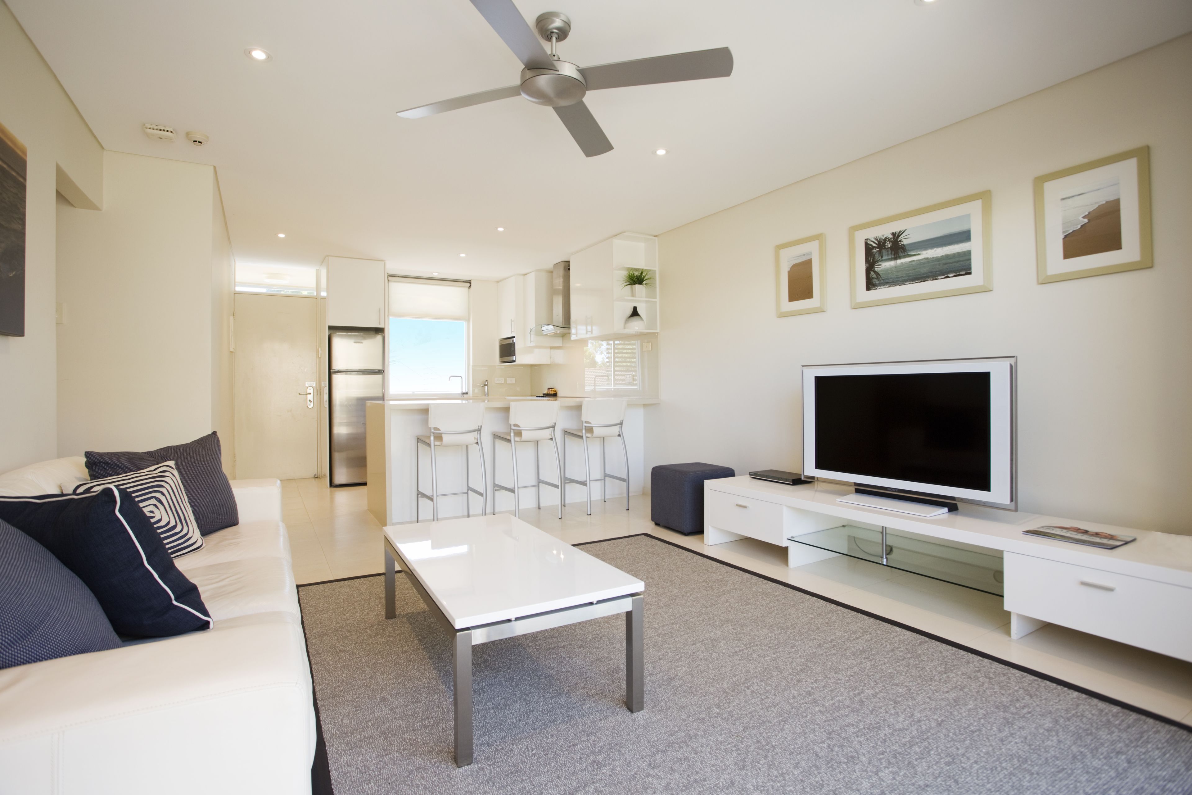 Un salon moderne avec un canapé blanc, une table basse, un téléviseur posé sur un meuble blanc, un ventilateur de plafond et, au fond, un coin cuisine.