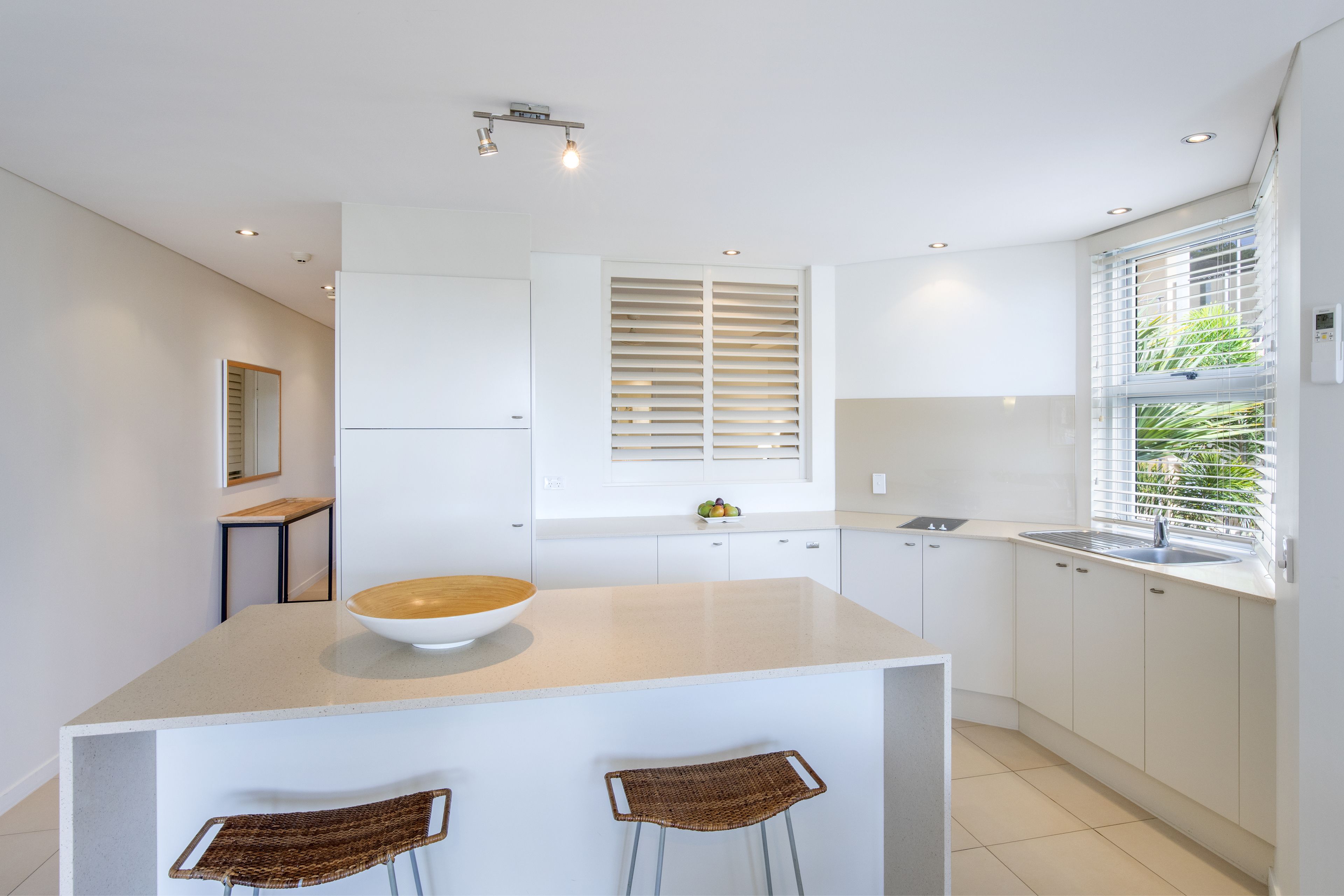 A modern kitchen with white cabinetry, a central island with a bowl, two wicker chairs, a window with blinds, and a small table with fruit.