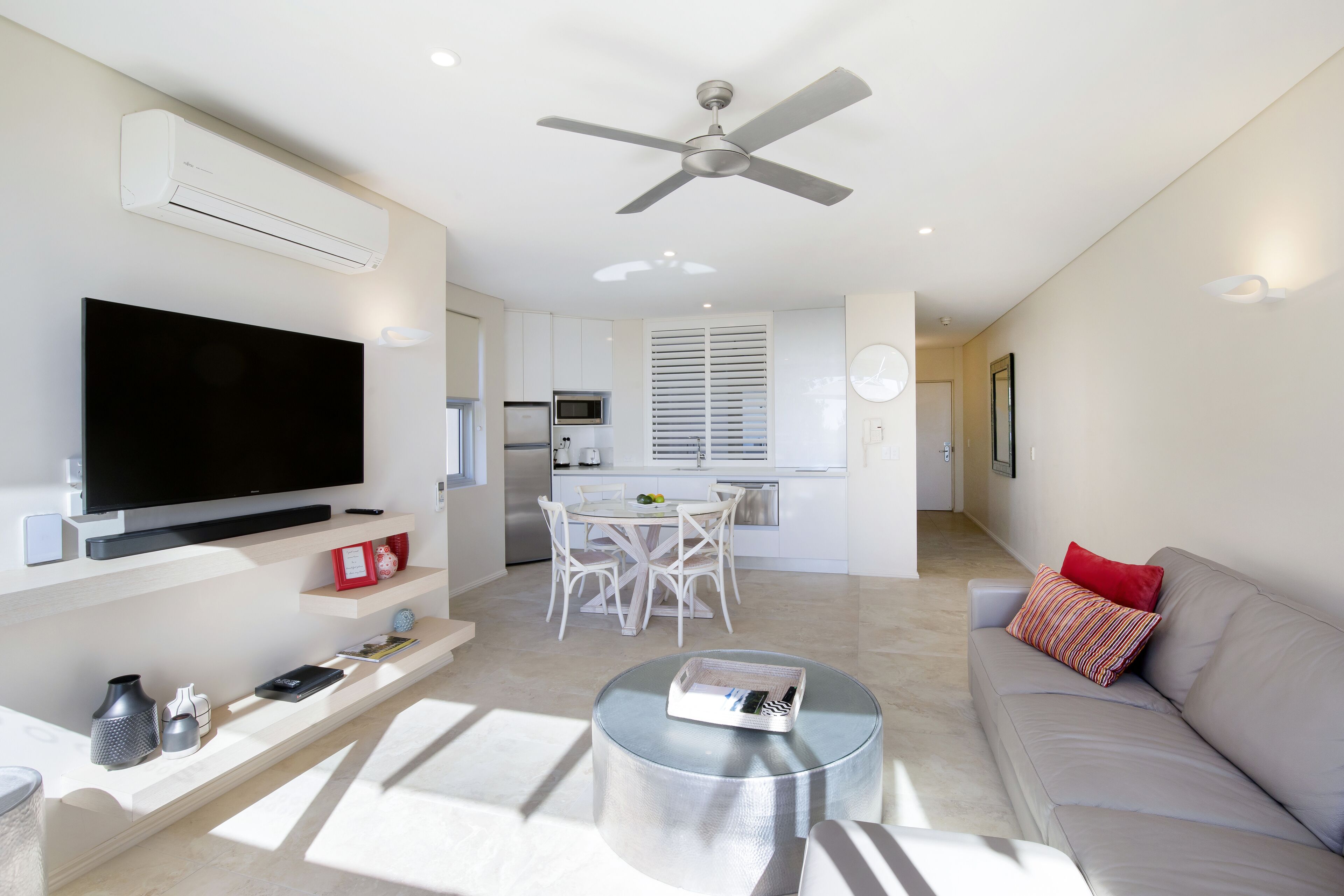 A modern living room with a flat-screen TV, a ceiling fan, a dining area with a white table and chairs, and a grey sofa with a red pillow.