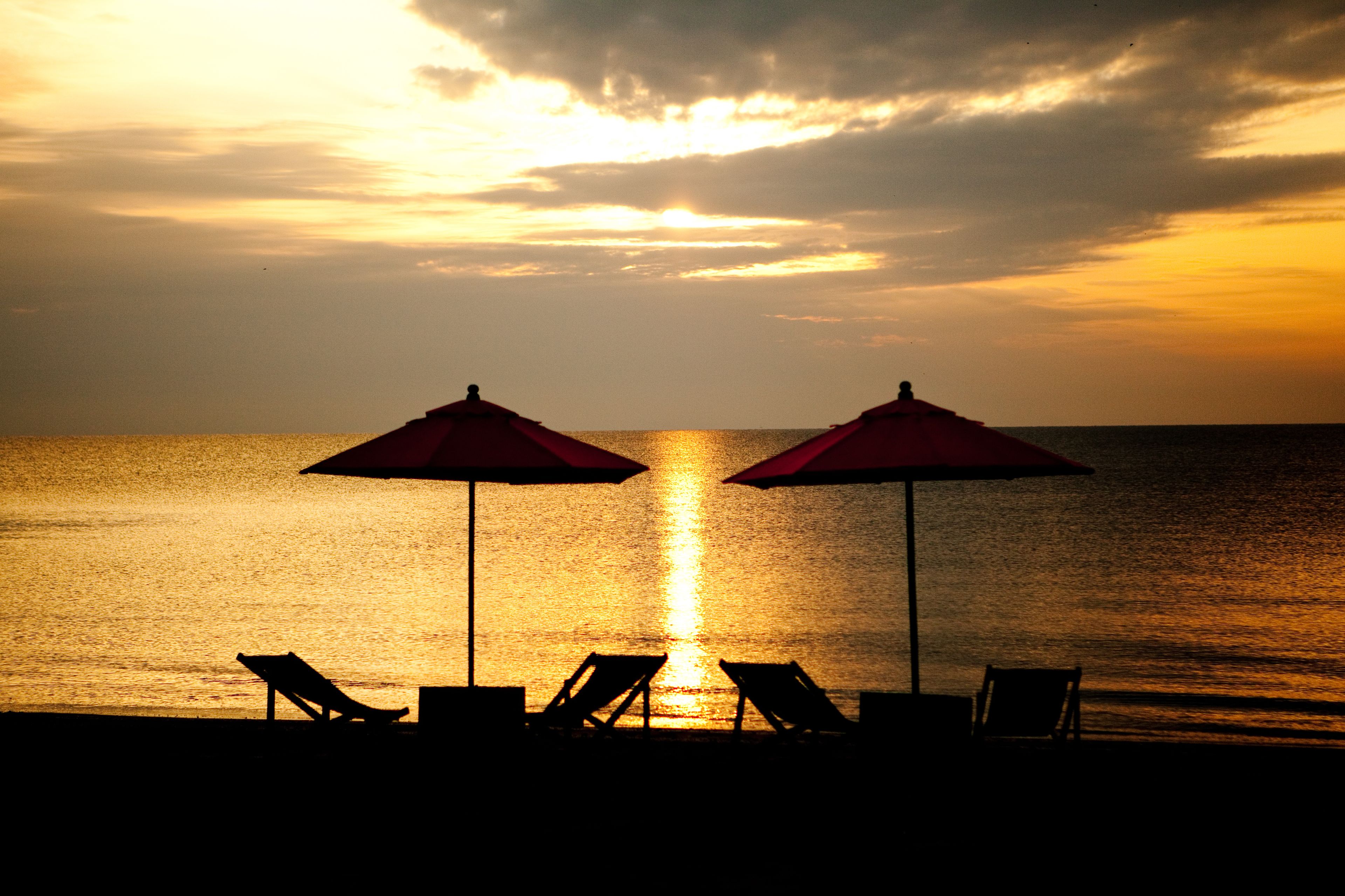 A beach scene with two umbrellas and lounge chairs facing the ocean at sunset.