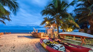 A beach with kayaks and a boat, palm trees, and a wooden structure with lights. - Vartika Resovilla KuiBuri Beach Resort and Villas (Prachuap Khiri Khan)